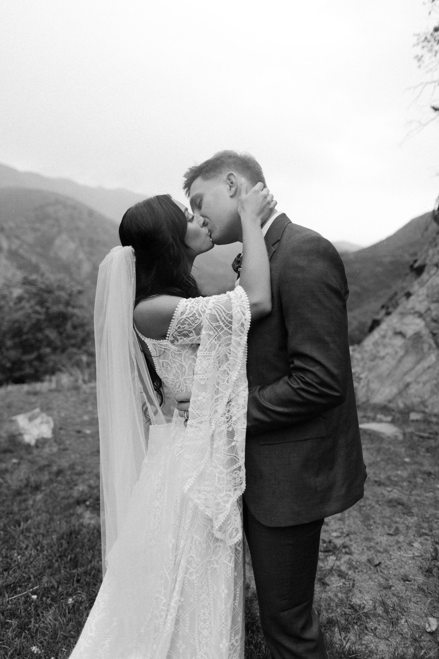 Bride and groom sharing a kiss in front of mountain scenery, bride wearing lace wedding dress with veil.
