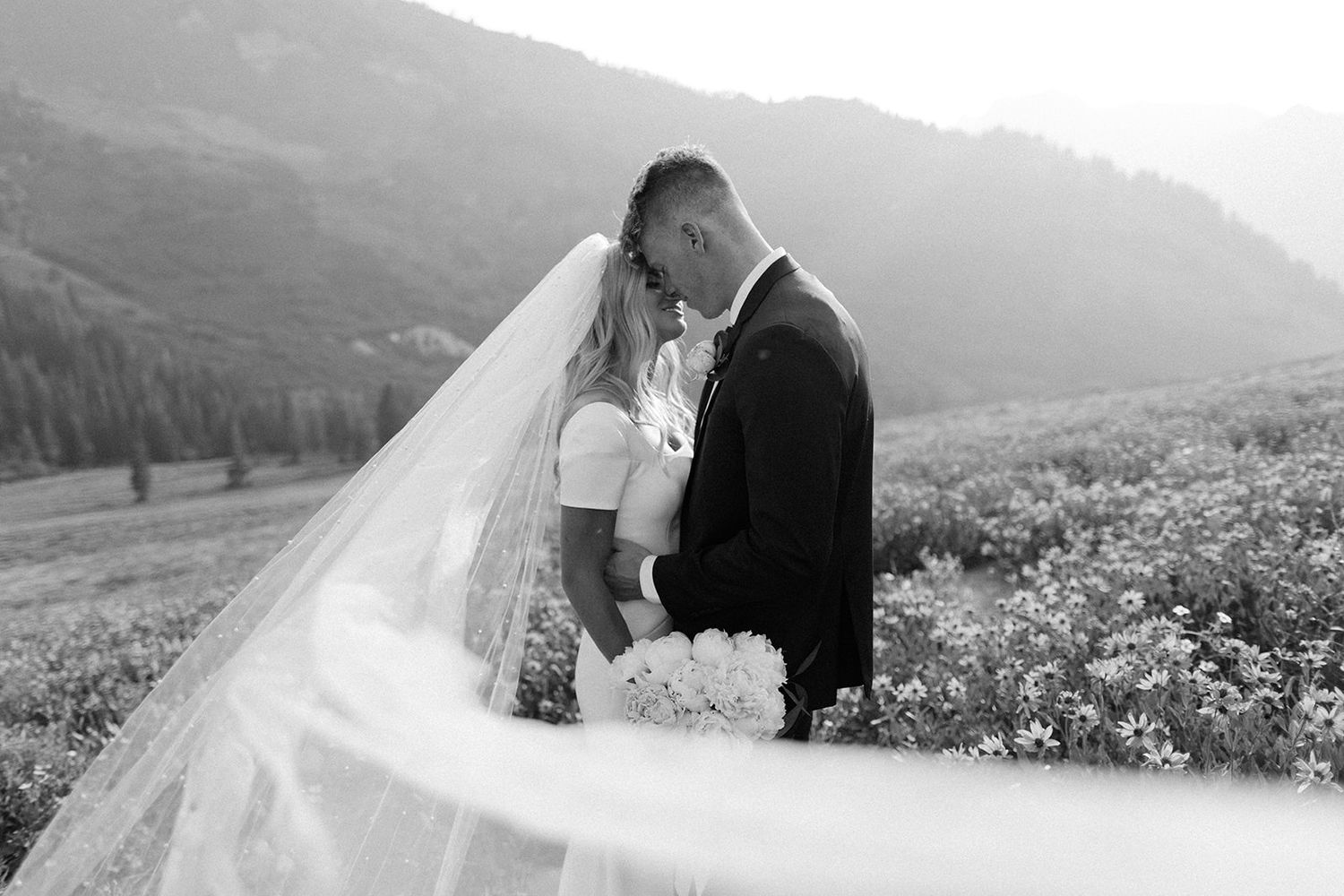 Black and white wedding photo of bride and groom embracing in mountain meadow with veil flowing.