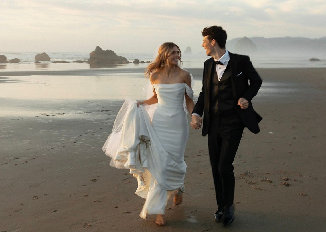 Bride and groom running joyfully along a dramatic beach shoreline, bride in off-shoulder gown.