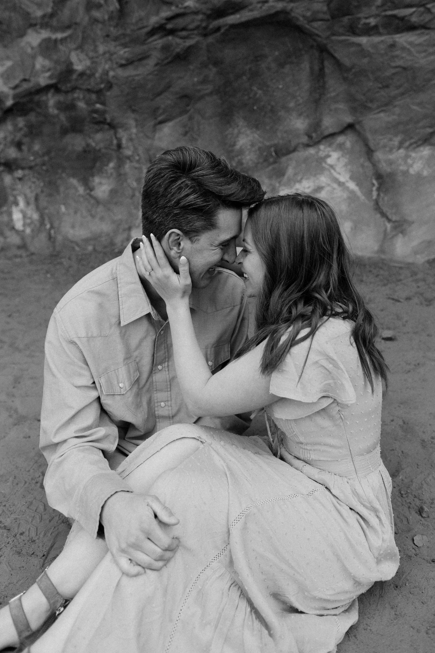 Black and white photo of couple sitting close together about to kiss near rocky background.