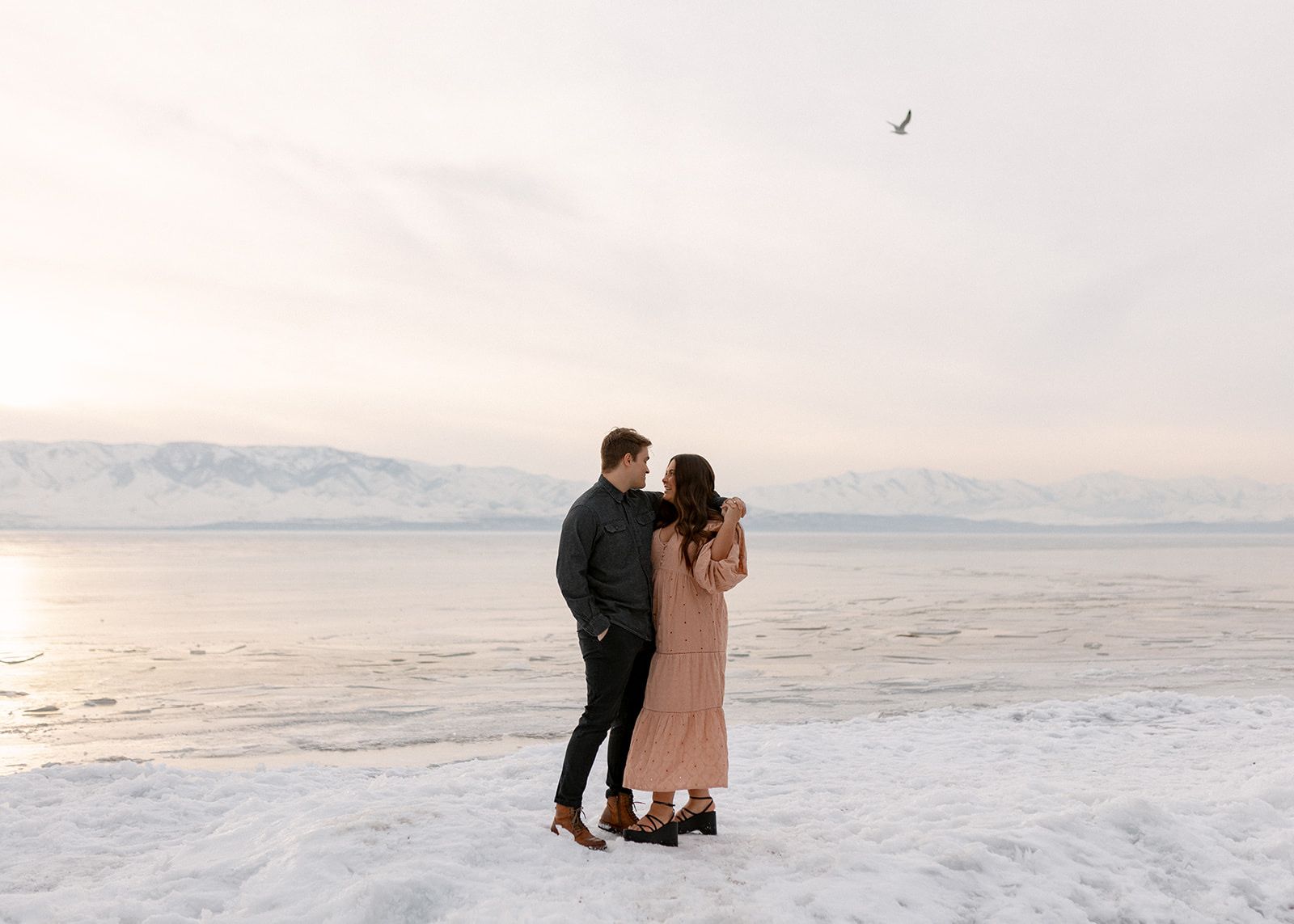 A romantic couple standing together on a snowy beach at sunset with mountain views.