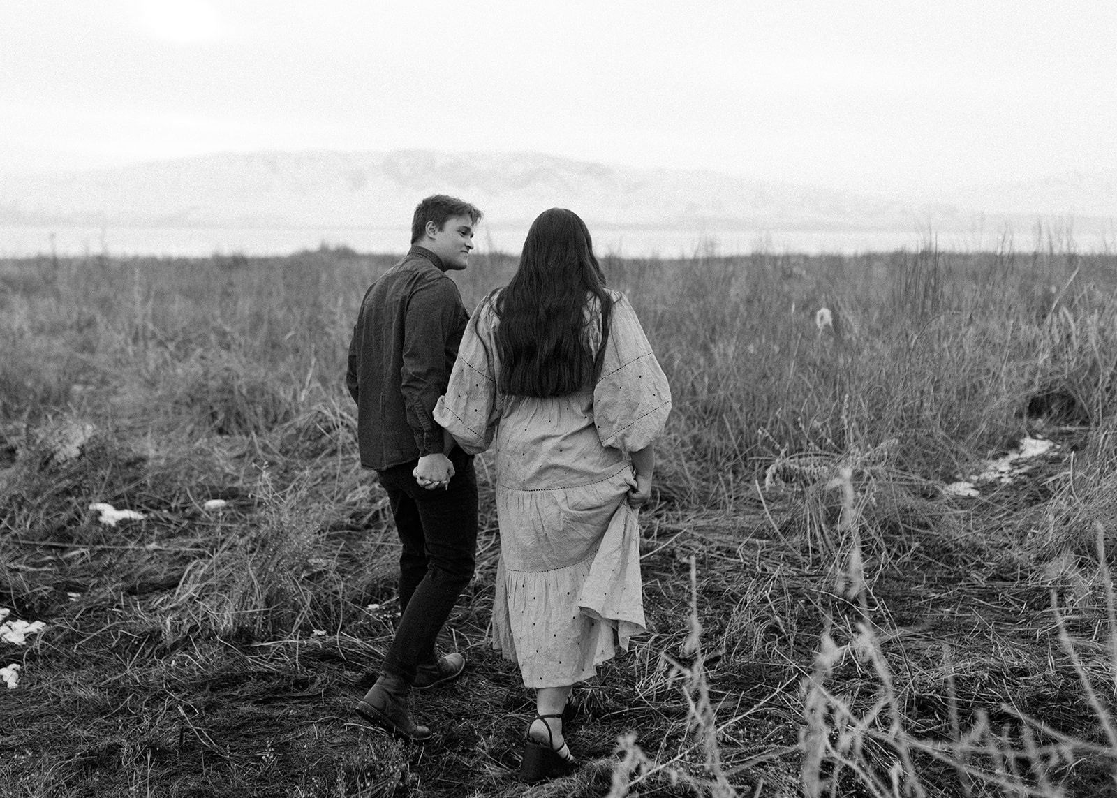 Black and white photo of couple walking hand in hand through a wild field with mountains.