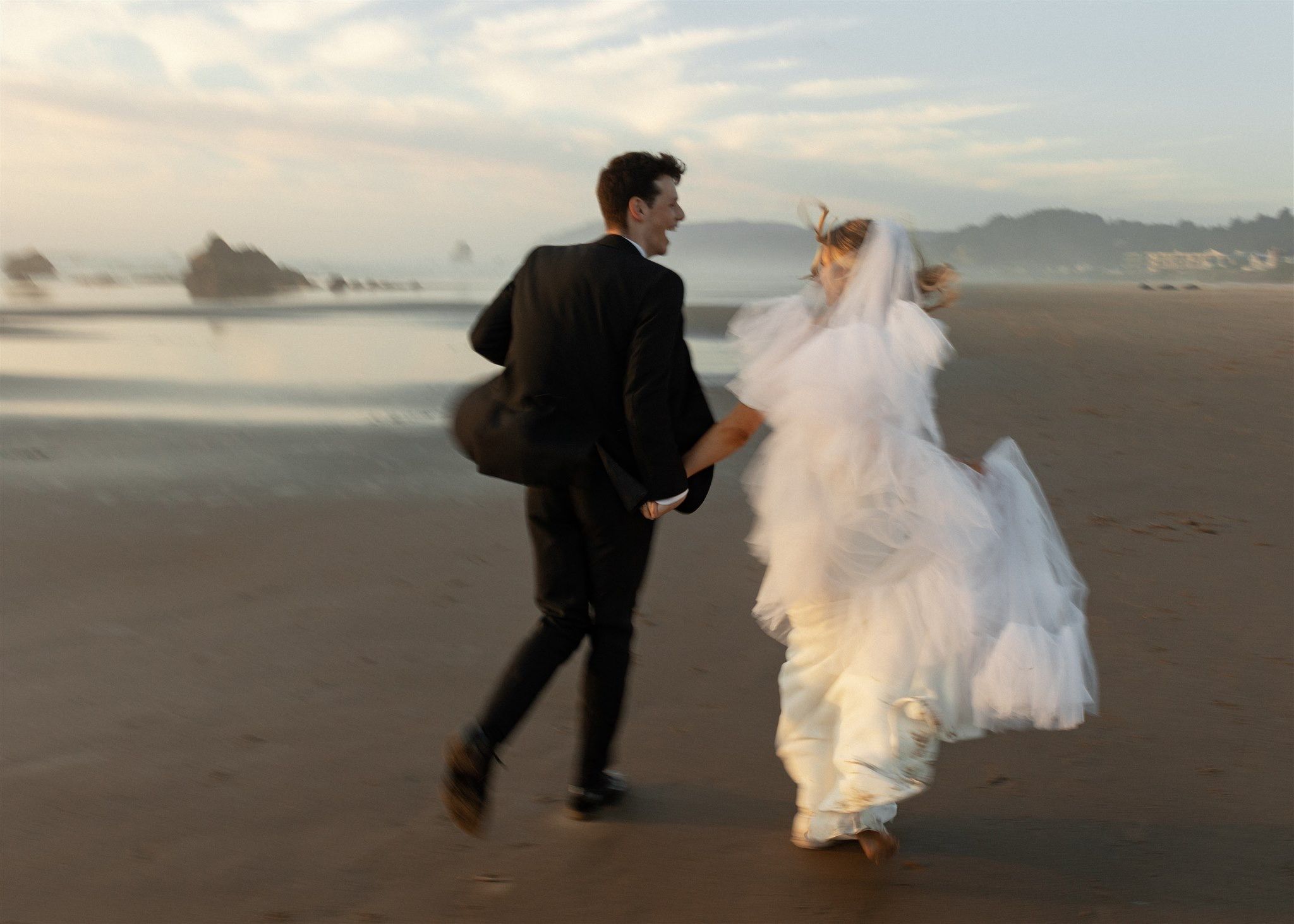 A bride and groom running joyfully along a scenic beach at dusk in wedding attire.