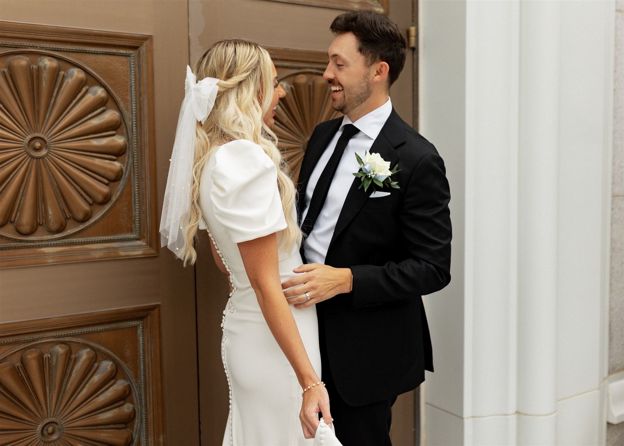 Bride and groom laughing together in front of ornate wooden door, she in white gown.