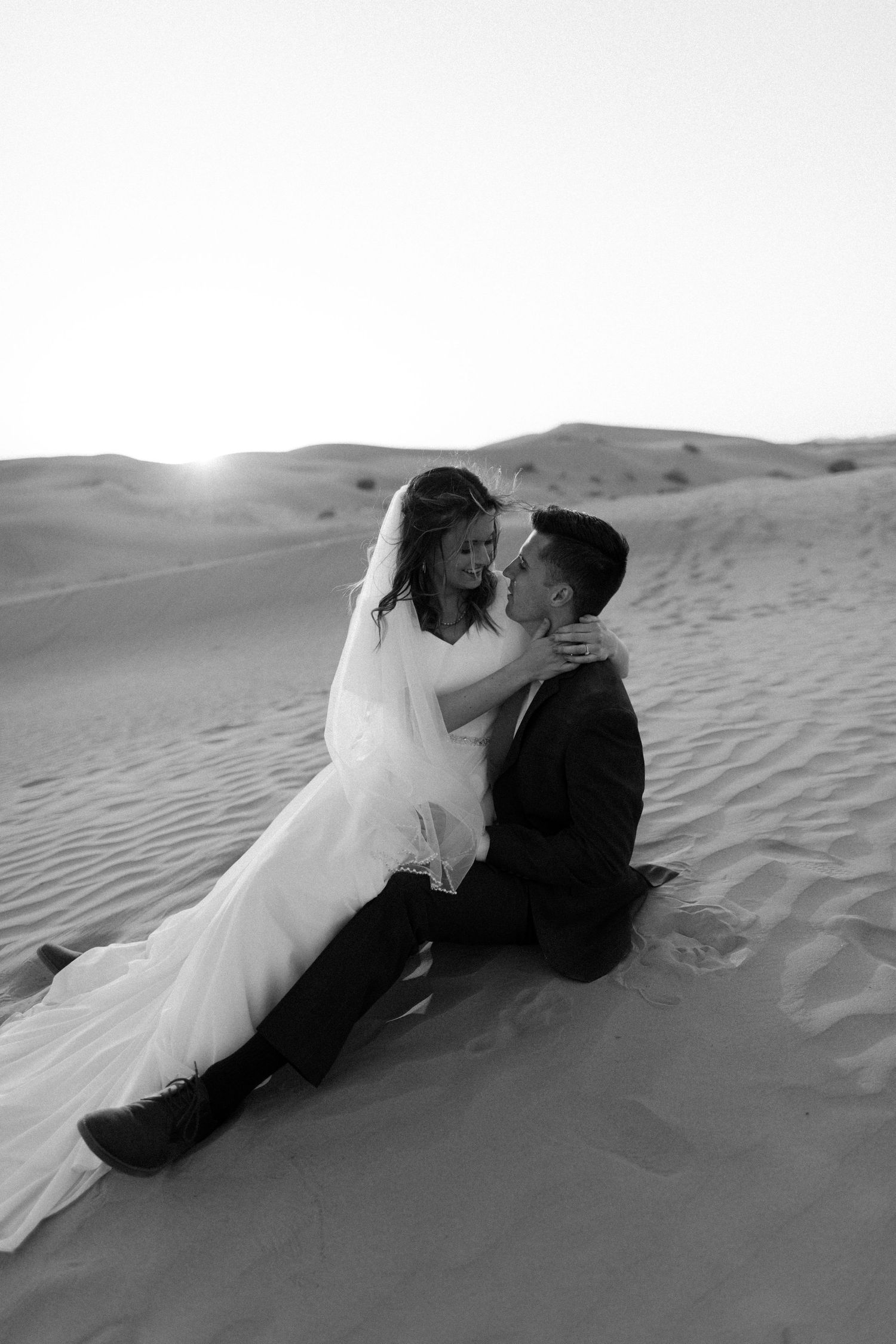Bride and groom sitting together on desert sand dunes at sunset in a romantic black and white photo.