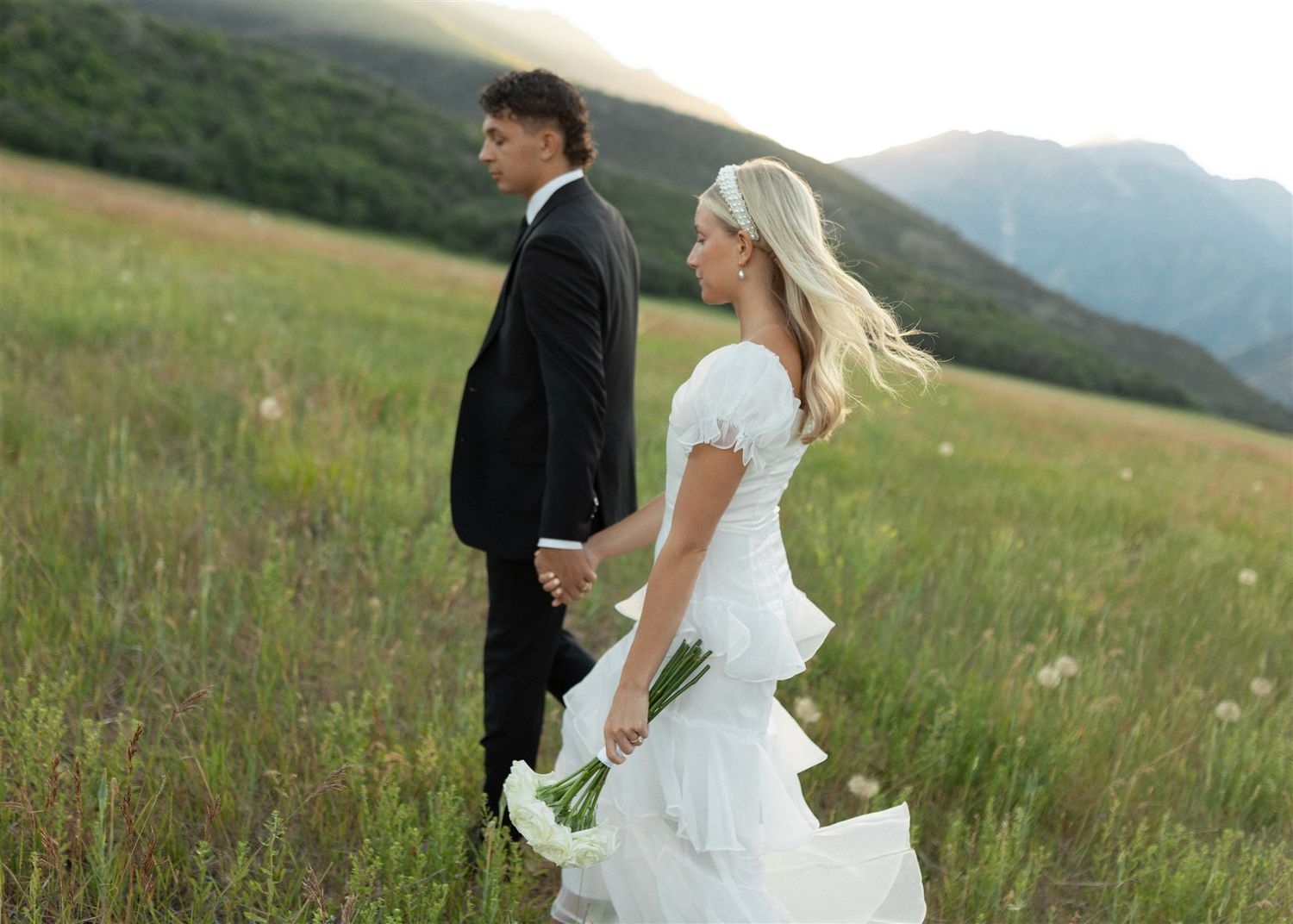 Bride in white ruffled gown and groom in black suit walk hand in hand through a green mountain meadow.