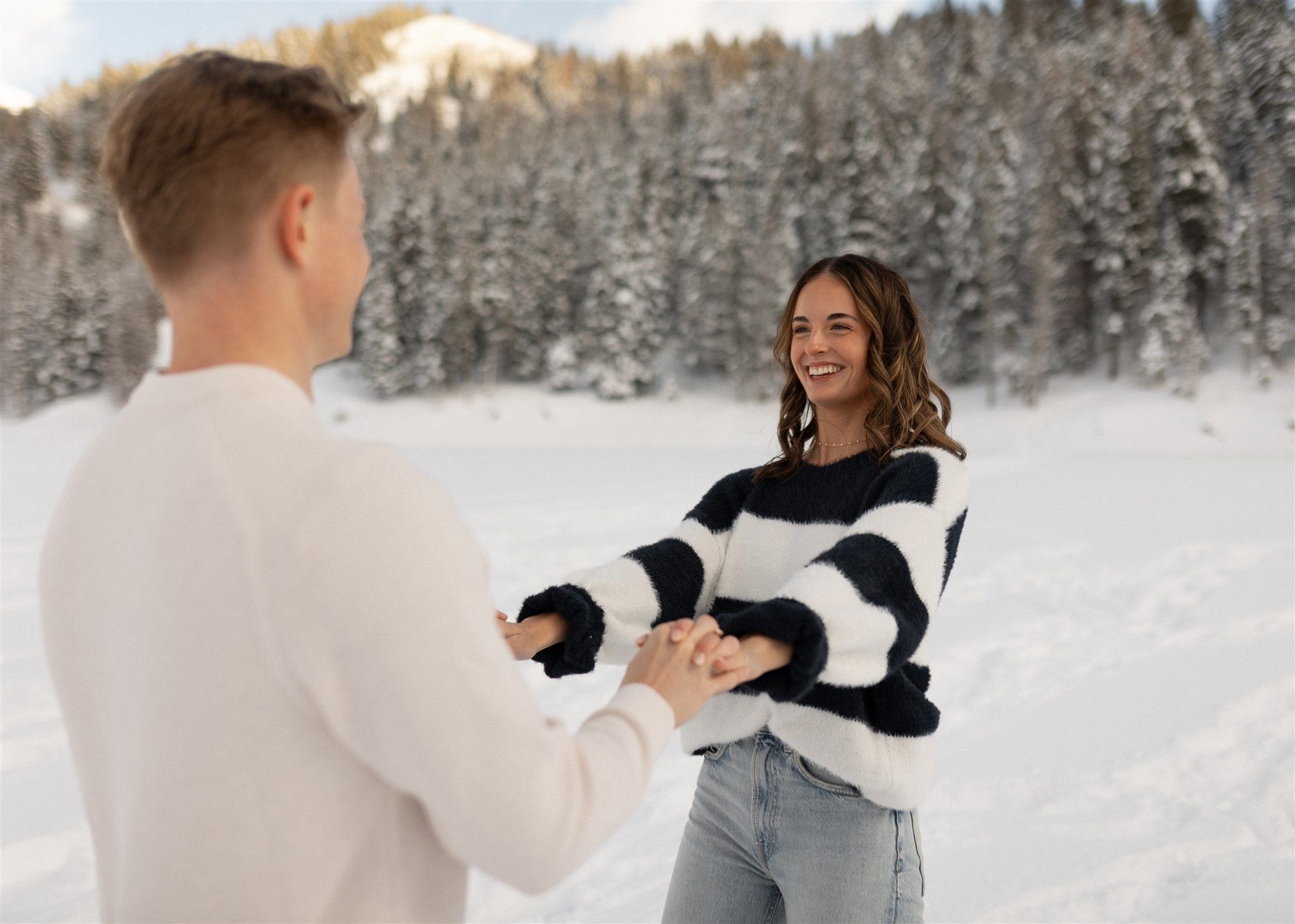 Couple holding hands and smiling in a snowy winter landscape with snow-covered pine trees.