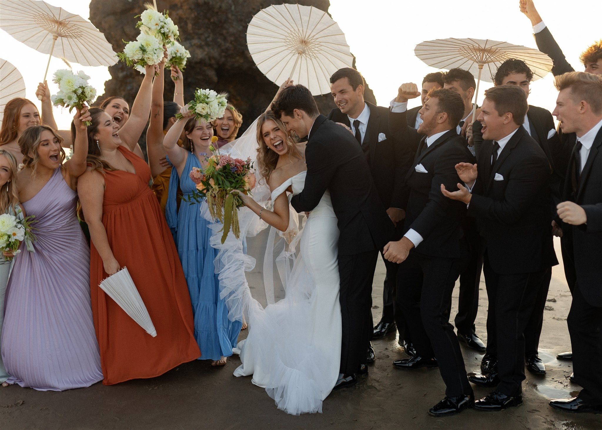 A joyful wedding party celebrating on a beach with colorful bridesmaid dresses and parasols.